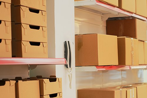 Empty warehouse with landline phone on wall, shelves and racks filled with merchandise in cardboard boxes. Multiple products prepared for shipment and transportation, stock goods.